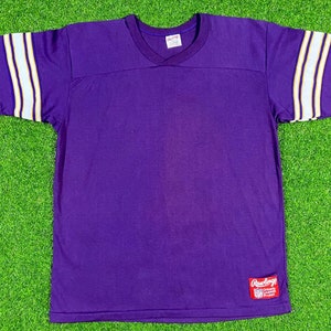May include: Purple short-sleeved jersey with white and gold stripes on the sleeves. The jersey has a v-neck and a Rawlings logo on the bottom right.