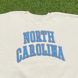 May include: Cream-colored sweatshirt with the words "NORTH CAROLINA" in light blue block letters. The text is arched across the chest. The sweatshirt has a crew neck and a simple, classic design.