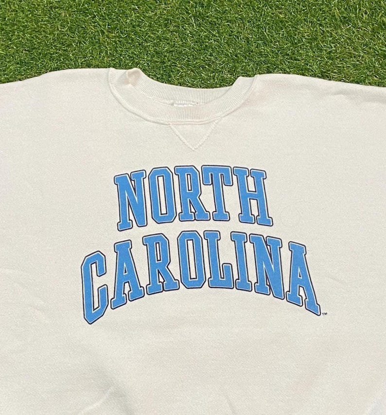 May include: Cream-colored sweatshirt with the words "NORTH CAROLINA" in light blue block letters. The text is arched across the chest. The sweatshirt has a crew neck and a simple, classic design.