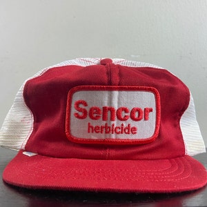 Puede incluir: Un gorro de camionero rojo y blanco con un parche blanco que dice "Sencor herbicide" en letras rojas.