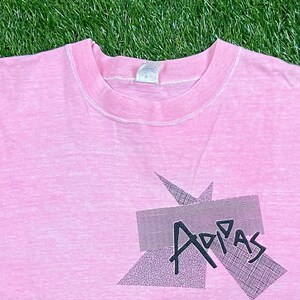 Peut inclure: Un t-shirt rose avec un motif graphique noir qui dit "Adidas" dans une police de caract&egrave;res stylis&eacute;e.