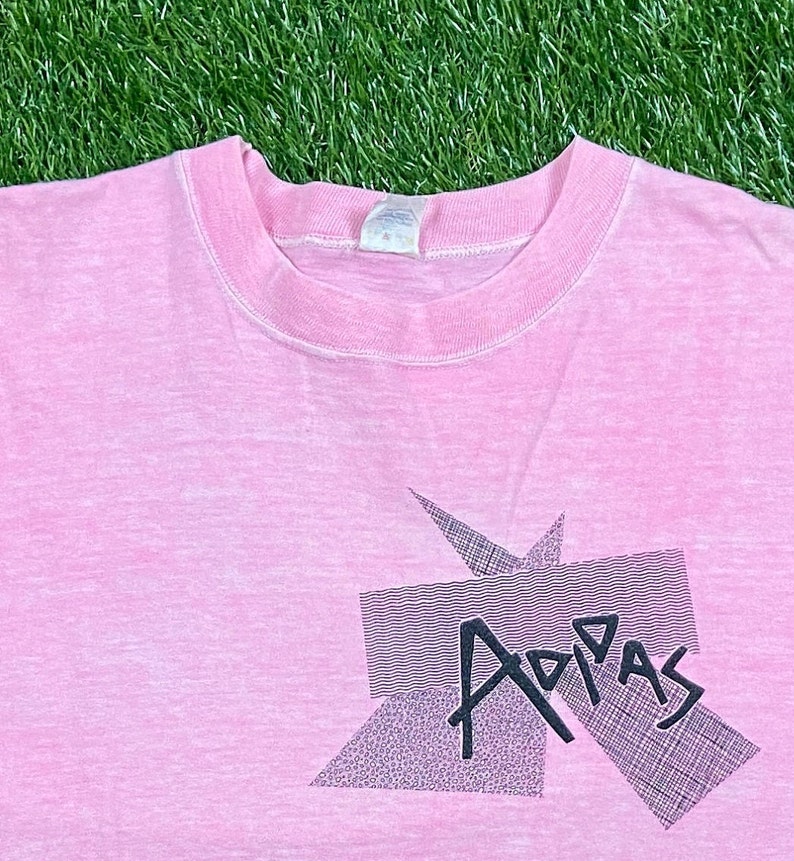 Peut inclure: Un t-shirt rose avec un motif graphique noir qui dit "Adidas" dans une police de caract&egrave;res stylis&eacute;e.