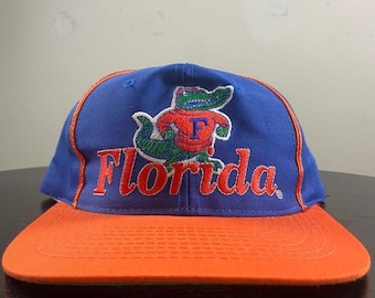 May include: A blue and orange baseball cap with an embroidered alligator wearing a jersey with the letter "F" and the word "Florida" on the front.