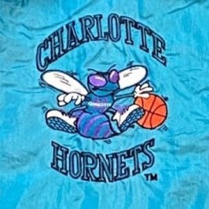 Może przedstawiać: Naszywka haftowana w kolorze niebieskim i białym z logo Charlotte Hornets. Logo przedstawia kresk&oacute;wkowego szerszenia z skrzydłami, trzymającego piłkę do koszyk&oacute;wki. Tekst "Charlotte Hornets" znajduje się nad szerszeniem, a "TM" pod nim.