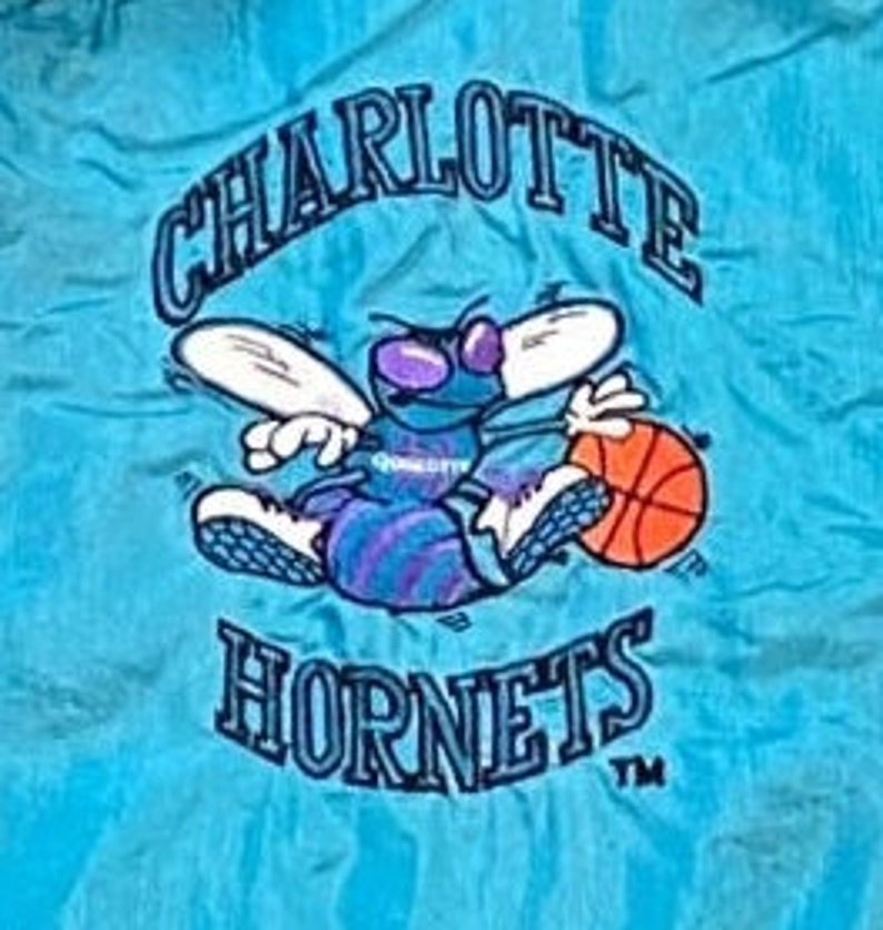 Może przedstawiać: Naszywka haftowana w kolorze niebieskim i białym z logo Charlotte Hornets. Logo przedstawia kresk&oacute;wkowego szerszenia z skrzydłami, trzymającego piłkę do koszyk&oacute;wki. Tekst "Charlotte Hornets" znajduje się nad szerszeniem, a "TM" pod nim.
