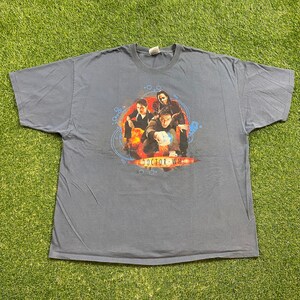 May include: A blue t-shirt with a graphic of the Doctor Who characters, including the Tenth Doctor, Rose Tyler, and Captain Jack Harkness. The graphic is surrounded by a blue and white circle with the text "Doctor Who" in white.