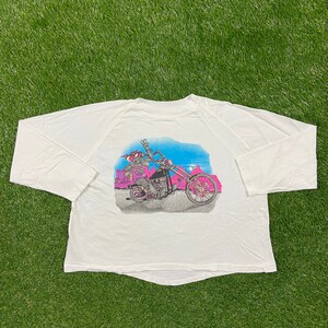 Peut inclure: T-shirt à manches longues blanche avec un personnage de dessin animé rose conduisant une moto. Le personnage porte un casque et fait un signe de paix. La moto est devant un ciel bleu avec des nuages et un paysage désertique.