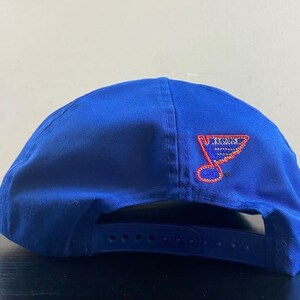 K&ouml;nnte beinhalten: Eine blaue Baseballkappe mit einem roten und wei&szlig;en gestickten St. Louis Blues Logo auf der R&uuml;ckseite.