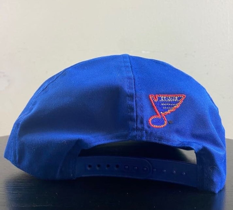 K&ouml;nnte beinhalten: Eine blaue Baseballkappe mit einem roten und wei&szlig;en gestickten St. Louis Blues Logo auf der R&uuml;ckseite.