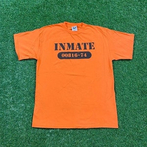 May include: Orange t-shirt with black text that reads "INMATE 00816-74".