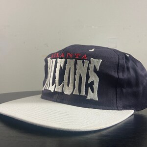 May include: A black and white baseball cap with the words "Atlanta Falcons" embroidered on the front in white.