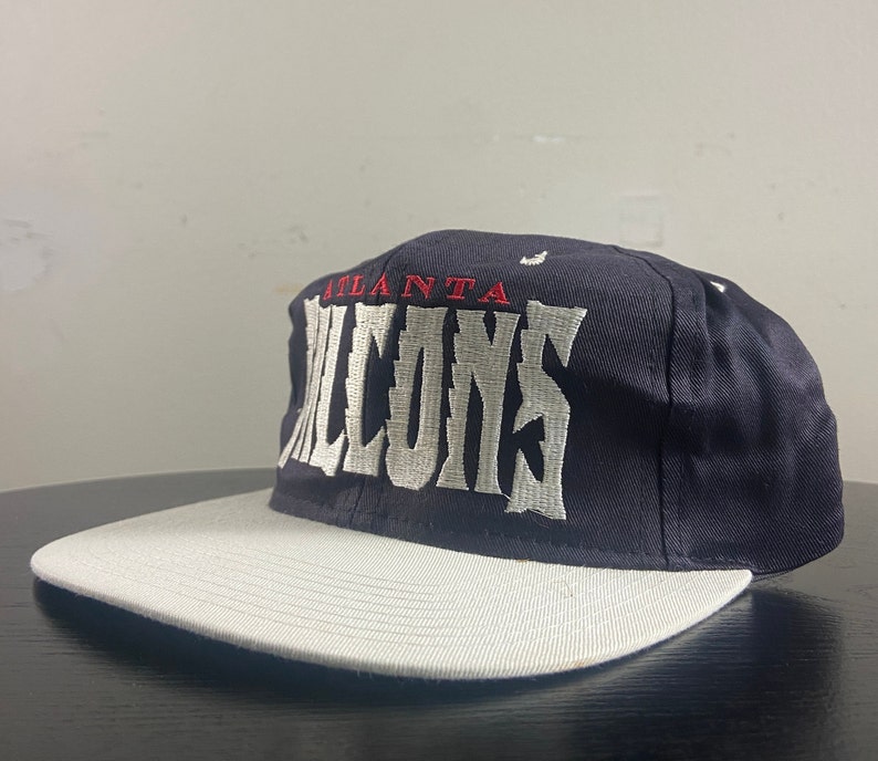 May include: A black and white baseball cap with the words "Atlanta Falcons" embroidered on the front in white.