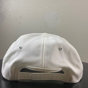 May include: A white baseball cap with a fabric strap and two silver eyelets on the back.
