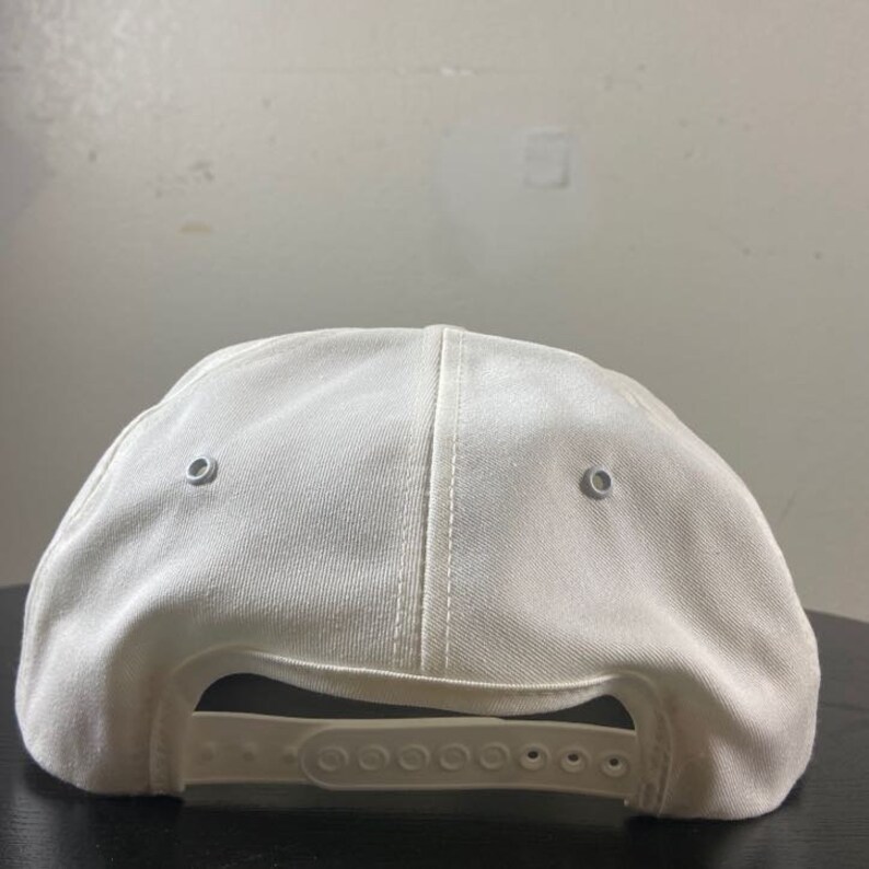 May include: A white baseball cap with a fabric strap and two silver eyelets on the back.