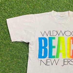 May include: White t-shirt with the words "WILDWOOD BEACH NEW JERS" printed in a colorful gradient. The word "BEACH" is in a rainbow gradient of blue, green, yellow, and orange. The shirt is on a green grassy surface.