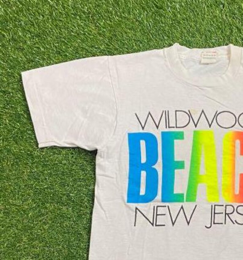 May include: White t-shirt with the words "WILDWOOD BEACH NEW JERS" printed in a colorful gradient. The word "BEACH" is in a rainbow gradient of blue, green, yellow, and orange. The shirt is on a green grassy surface.