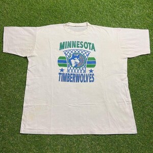 May include: Off-white vintage t-shirt featuring the Minnesota Timberwolves logo. The design includes the team name, a basketball hoop, a wolf graphic, and stars in green and blue. The shirt is made of a soft, worn fabric.