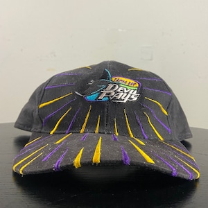 May include: Black baseball cap with a yellow and purple sunburst design. The front of the cap features an embroidered logo that says "Tampa Bay Devil Rays".