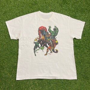 Op de afbeelding: Wit T-shirt met een grafische print van een groep superhelden, waaronder Thanos, Hulk en anderen. De grafiek is in een stripboek stijl en gebruikt felle kleuren.