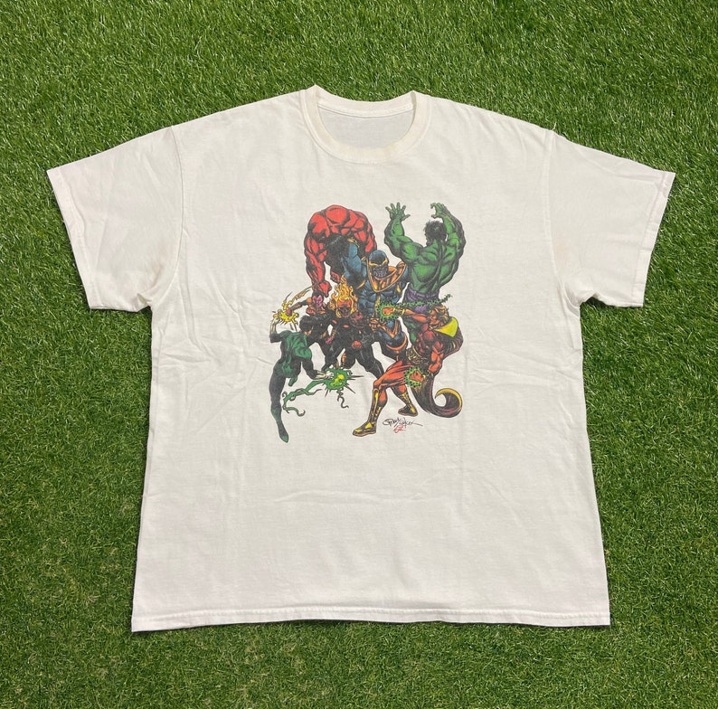 Op de afbeelding: Wit T-shirt met een grafische print van een groep superhelden, waaronder Thanos, Hulk en anderen. De grafiek is in een stripboek stijl en gebruikt felle kleuren.