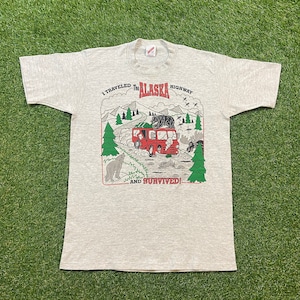 May include: A grey t-shirt featuring a graphic print of a red truck driving through a forest with a bear, moose, and wolves. The text on the shirt reads "I travelled the Alaska Highway and survived!"