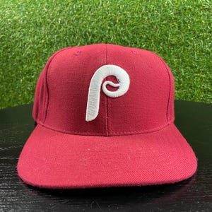 Può includere: Un cappellino da baseball color bordeaux a tinta unita con una lettera P bianca ricamata in rilievo sul pannello frontale. Il cappellino presenta una corona strutturata e una visiera piatta, con cuciture abbinate e occhielli di ventilazione.
