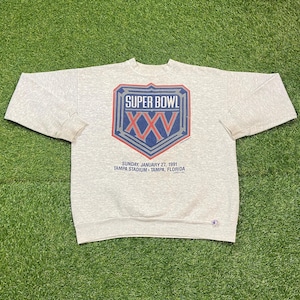May include: A light gray Super Bowl XXV sweatshirt. The front features a shield-shaped graphic with "SUPER BOWL" at the top and "XXV" in red and blue. Below, text reads "SUNDAY JANUARY 27, 1991 TAMPA STADIUM - TAMPA, FLORIDA."