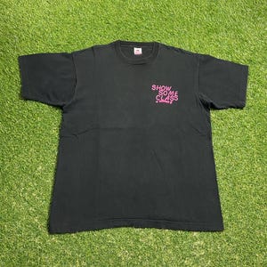 May include: Black t-shirt with the text "SHOW SOME CLASS" in pink. The shirt is a crew neck style and appears to be made of cotton. The t-shirt is laid flat on a green grassy surface.