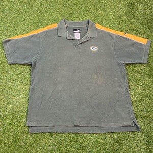 Puede incluir: Polo verde con rayas amarillas en los hombros y el logo de los Green Bay Packers. La camisa tiene cuello y mangas cortas. El logo de Puma es visible en el cuello.