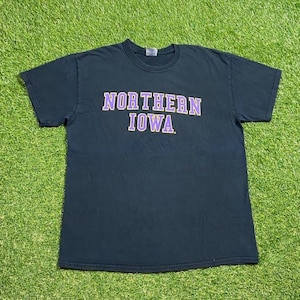 May include: Black t-shirt featuring the text "NORTHERN IOWA" in purple lettering. The crew neck shirt is made of cotton. The t-shirt is displayed on a green surface.
