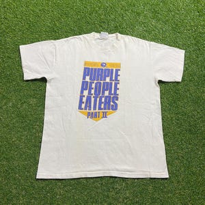 May include: White t-shirt with the text "PURPLE PEOPLE EATERS PART II" in blue and yellow. The shirt also has the words "MINNESOTA VIKINGS" at the top. The shirt is laid flat on a green surface.