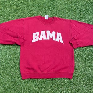 May include: A crimson sweatshirt with the word "BAMA" in white block letters across the chest. The long-sleeved crew neck top is made of a soft material and is laid out on a green surface.