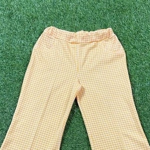 May include: A pair of yellow and white gingham patterned pants. The pants have an elastic waistband and a flared leg design. The fabric appears to be a lightweight material, suitable for warmer weather. The pants are displayed on a green surface.