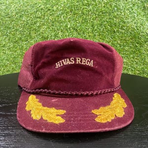 May include: A maroon velvet baseball cap featuring a braided cord trim above the brim and gold embroidered leaf designs on the visor. The front panel displays the text HIVAS REGA in gold embroidery, and the sides consist of matching mesh panels.
