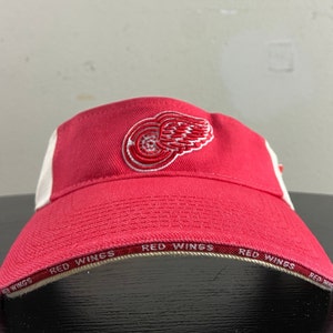 Peut inclure: Casquette de baseball rouge et blanche avec un logo rouge Detroit Red Wings sur le devant. Le bord de la casquette porte les mots "Red Wings" répétés tout autour.