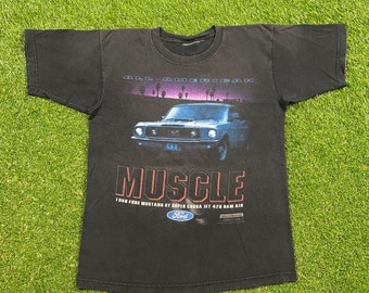Op de afbeelding: Zwart T-shirt met een afbeelding van een blauwe Ford Mustang Super Cobra Jet 428 Ram Air. De afbeelding bevat de tekst "Muscle" en "All American". Het Ford-logo is ook op het T-shirt te zien.