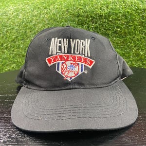 Puede incluir: Una gorra de béisbol negra con un logotipo bordado en la parte delantera. El texto NEW YORK aparece en blanco sobre una banda roja con la palabra YANKEES en blanco. Debajo de la banda hay una forma de home plate blanca con un gráfico de béisbol.