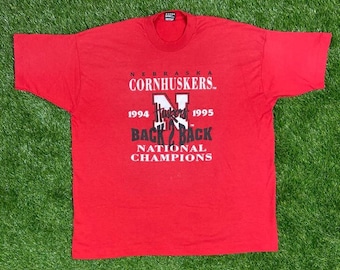 Könnte beinhalten: Rotes T-Shirt mit dem Text "Nebraska Cornhuskers" und "Back 2 Back National Champions" mit den Jahren 1994 und 1995.