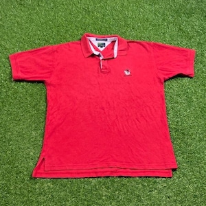 Puede incluir: Polo rojo de manga corta con cuello blanco y verde y un pequeño logotipo bordado en el pecho izquierdo. El logotipo es un bulldog rojo con la palabra "BULLDOG" debajo.