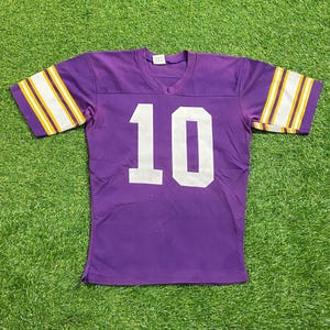 May include: A purple jersey with the number 10 in white on the front. The jersey has a V-neck and white and yellow stripes on the sleeves. The jersey is made of a soft, breathable material.