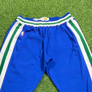 May include: Blue athletic pants with white and green stripes down the sides. The pants have a white waistband with green trim and a gold crown emblem. An NBA logo is visible on the left leg.