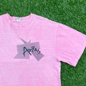 Peut inclure: Un t-shirt rose avec un aspect d&eacute;lav&eacute; et d&eacute;t&eacute;rior&eacute;. Le t-shirt pr&eacute;sente un motif graphique noir et blanc du mot "Adidas" dans un design g&eacute;om&eacute;trique.