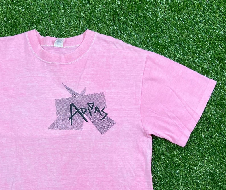 Peut inclure: Un t-shirt rose avec un aspect d&eacute;lav&eacute; et d&eacute;t&eacute;rior&eacute;. Le t-shirt pr&eacute;sente un motif graphique noir et blanc du mot "Adidas" dans un design g&eacute;om&eacute;trique.