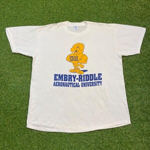 May include: White t-shirt featuring the Embry-Riddle Aeronautical University logo. The logo includes a yellow eagle mascot with the letters ERAU, and the university's name in blue text. The shirt is made of a soft material.