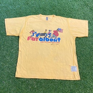 May include: Yellow short-sleeved t-shirt with a cartoon graphic of people pushing a shopping cart. The text "Fat Albert and the Junkyard Gang" is printed below the graphic.