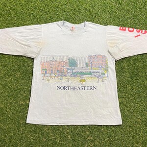 May include: White long-sleeve t-shirt with a colorful graphic of a city scene, including buildings, a trolley, and people. The word "NORTHEASTERN" is printed below the graphic. The right sleeve has the word "BOSTON" printed in red.
