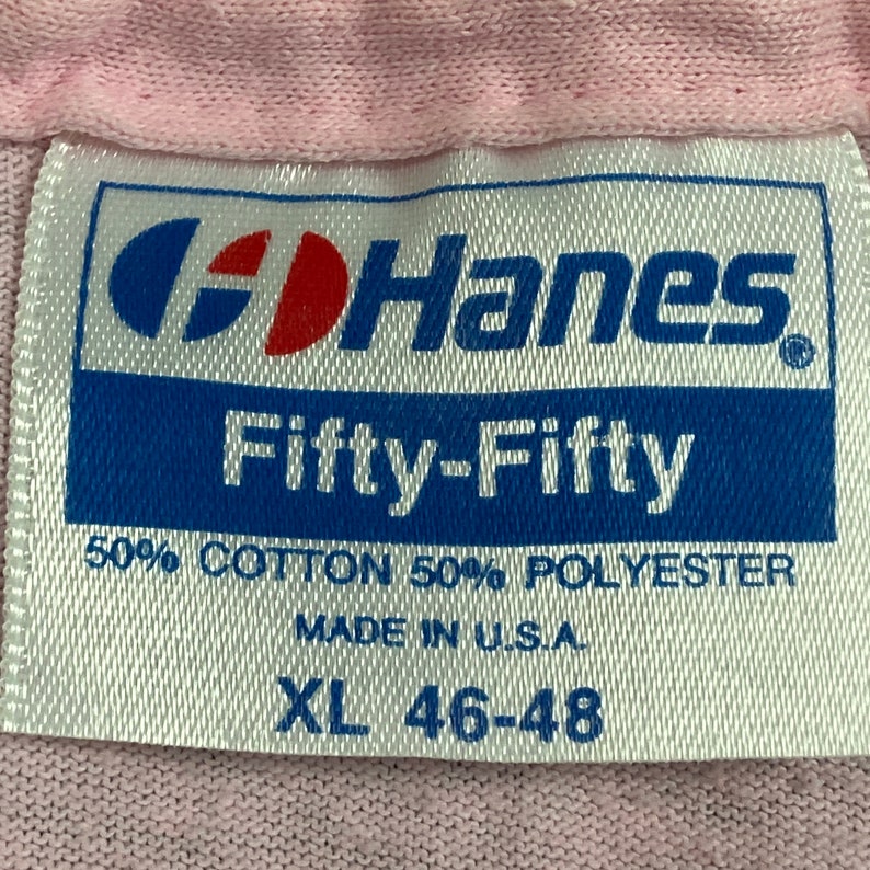 Puede incluir: Primer plano de una etiqueta Hanes Fifty-Fifty. La etiqueta es blanca con texto azul y rojo. Dice "Hanes Fifty-Fifty" y detalla la composici&oacute;n de la tela como 50% algod&oacute;n y 50% poli&eacute;ster. Talla XL 46-48. Hecho en EE. UU.