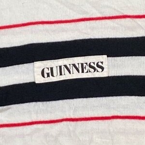 May include: A white striped shirt with a black and red stripe pattern. The shirt has a white label with the word "GUINNESS" printed in black.