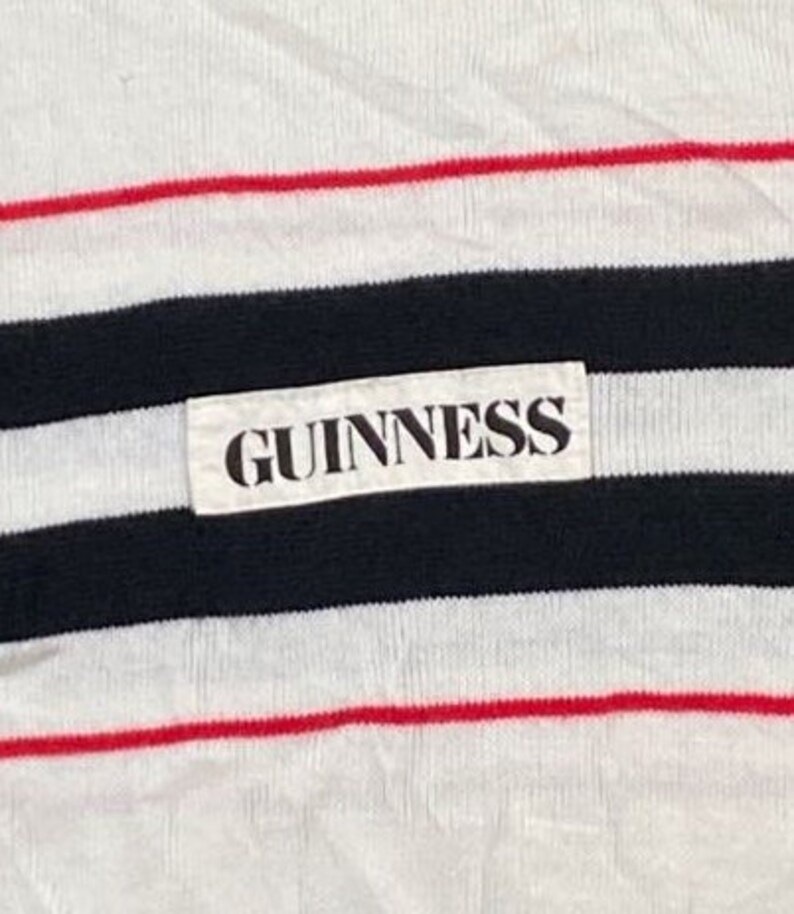 May include: A white striped shirt with a black and red stripe pattern. The shirt has a white label with the word "GUINNESS" printed in black.