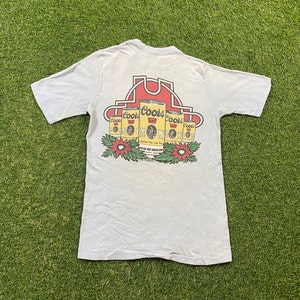 以下が含まれることがあります： 薄い青色のTシャツに、赤い白の馬蹄のデザインで円形に並んだ6つのクアーズライトビール缶のグラフィックがプリントされています。缶には「クアーズ」の文字が印刷されています。